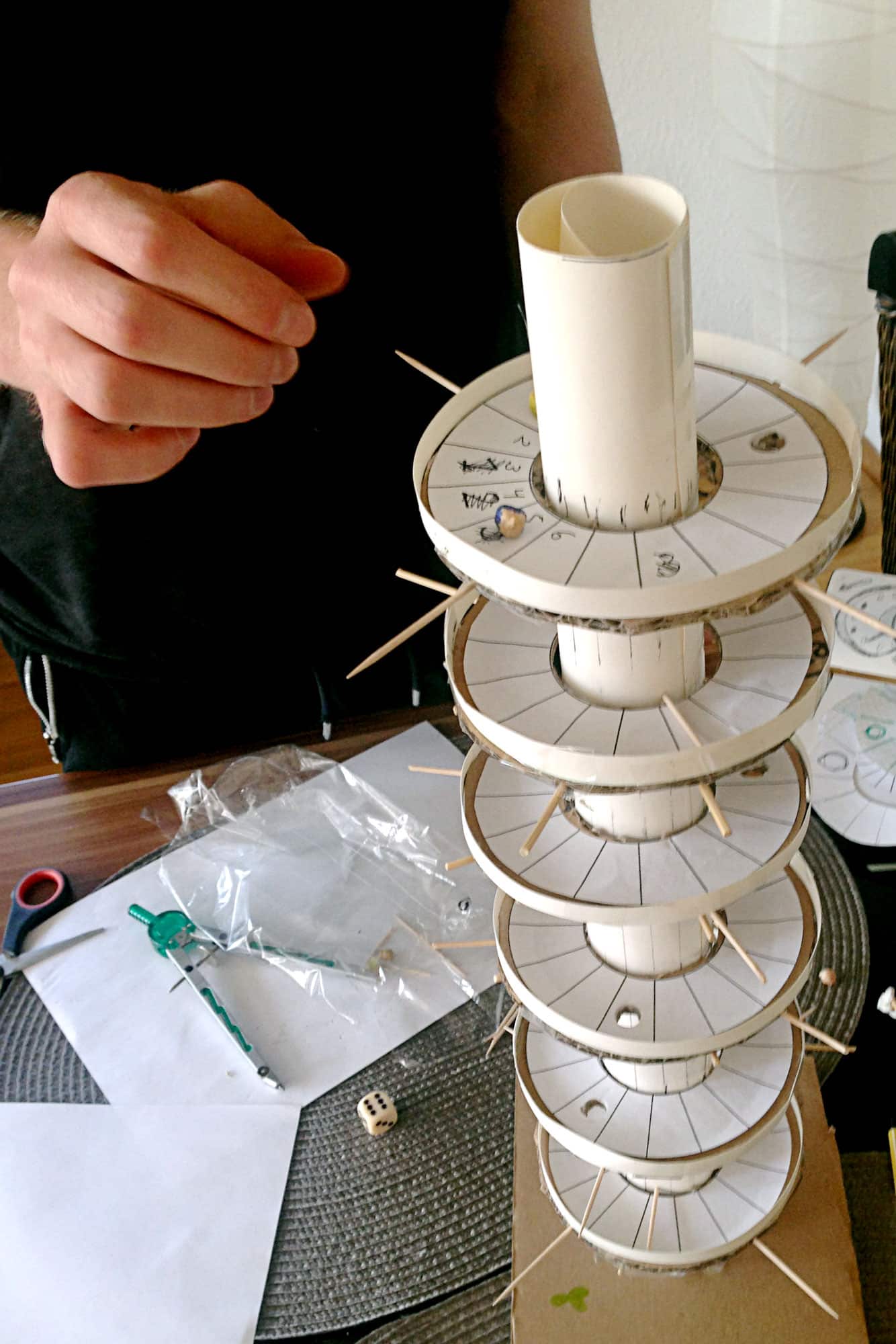 Person's hand assembling a tall stack of white plastic discs with pins on a central white tube, on a workbench with tools like compasses, scissors, paper, and a die nearby.